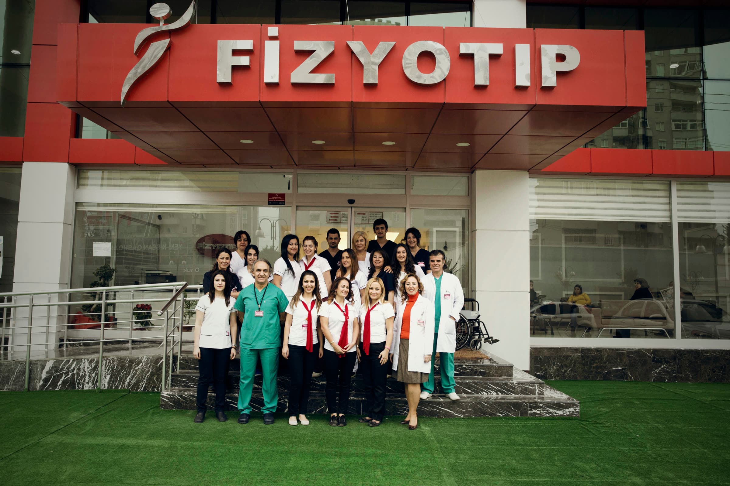 Fizyotıp ekibi, kırmızı Fizyotıp tabelalı merkez binasının önünde
